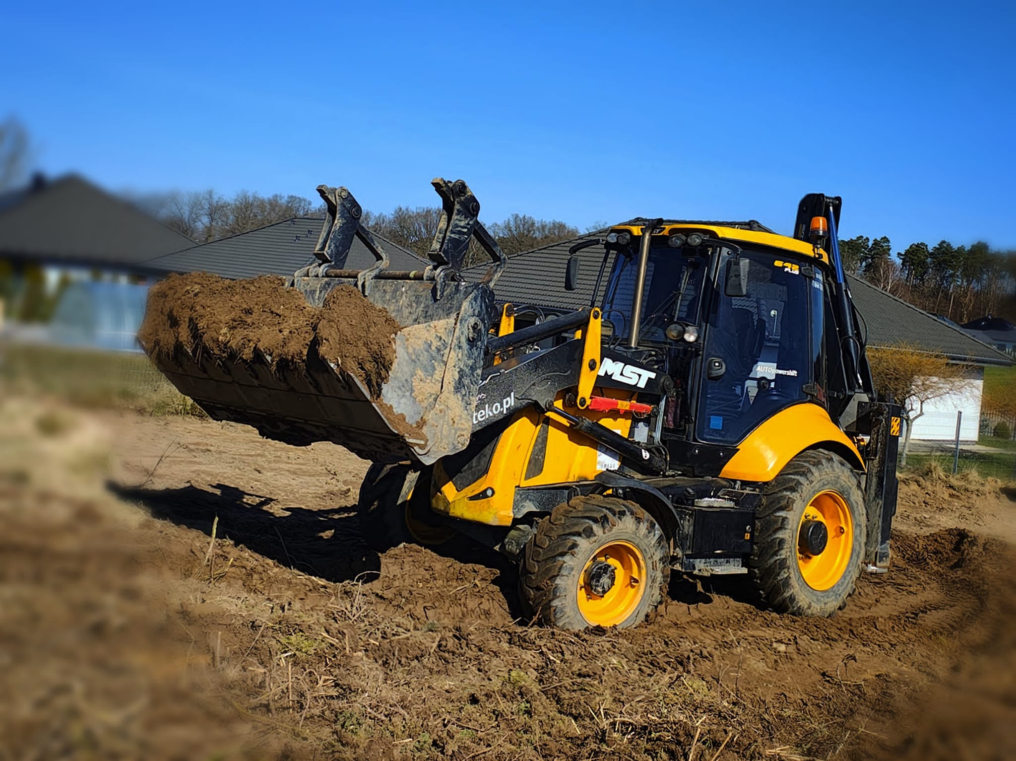 Żółto-czarna koparko-ładowarka MST z pełną łyżką ziemi na tle błękitnego nieba i zabudowań. Widoczne logo teko.pl na maszynie.