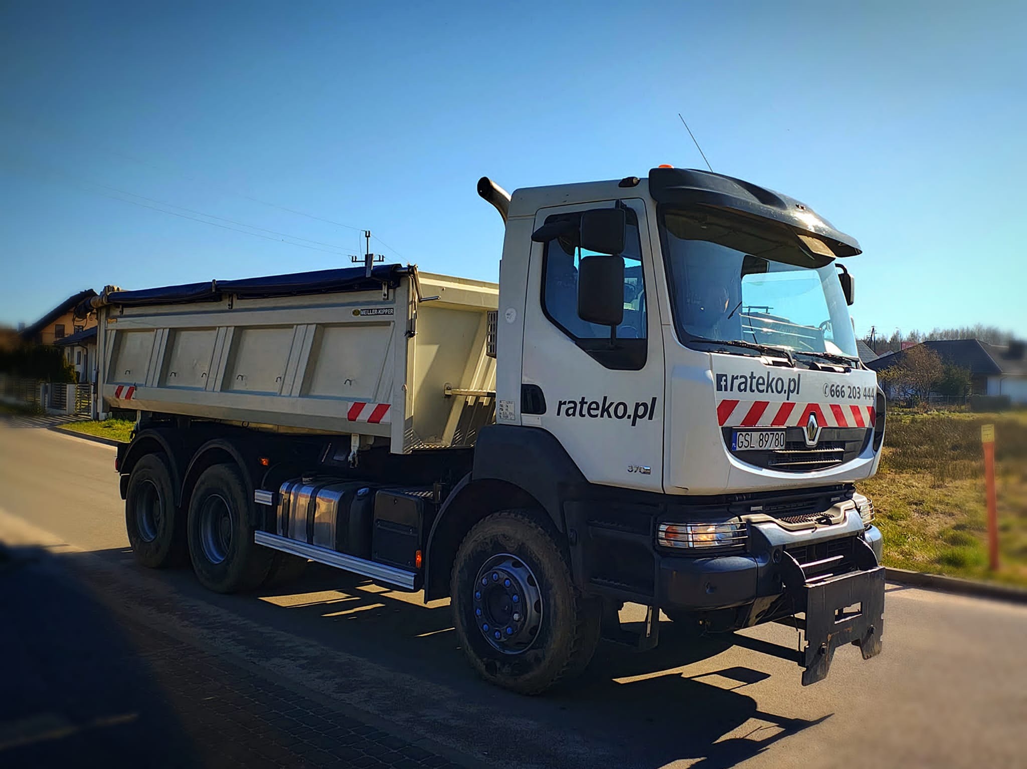 Ciężarówka wywrotka Renault z logo rateko.pl na boku, zaparkowana na drodze w słoneczny dzień. Widoczne detale kabiny, skrzyni ładunkowej i opon.