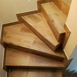 Schody wyłożone płytkami imitującymi drewno, z widocznymi fugami i listwami przypodłogowymi w kolorze płytek, sfotografowane z góry.