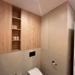 Nowoczesna łazienka w beżowych tonacjach z drewnianymi szafkami wiszącymi, białą toaletą podwieszaną i bidetem na tle płytek z geometrycznym wzorem.