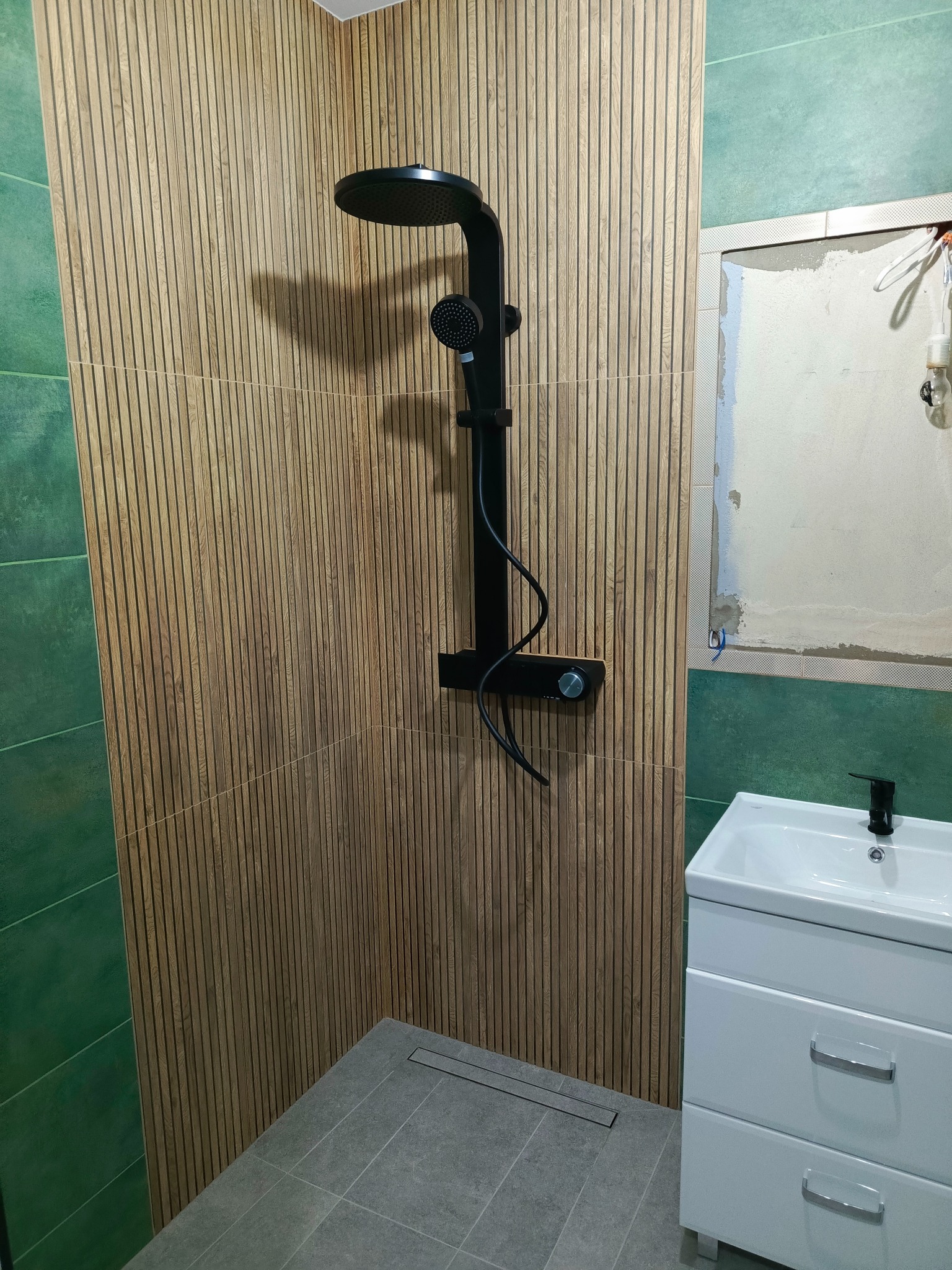 Nowoczesna łazienka z kabiną prysznicową typu walk-in, wykończona płytkami imitującymi drewniane lamele na ścianie, czarna armatura i biała szafka z umywalką, widoczne niedokończone prace...