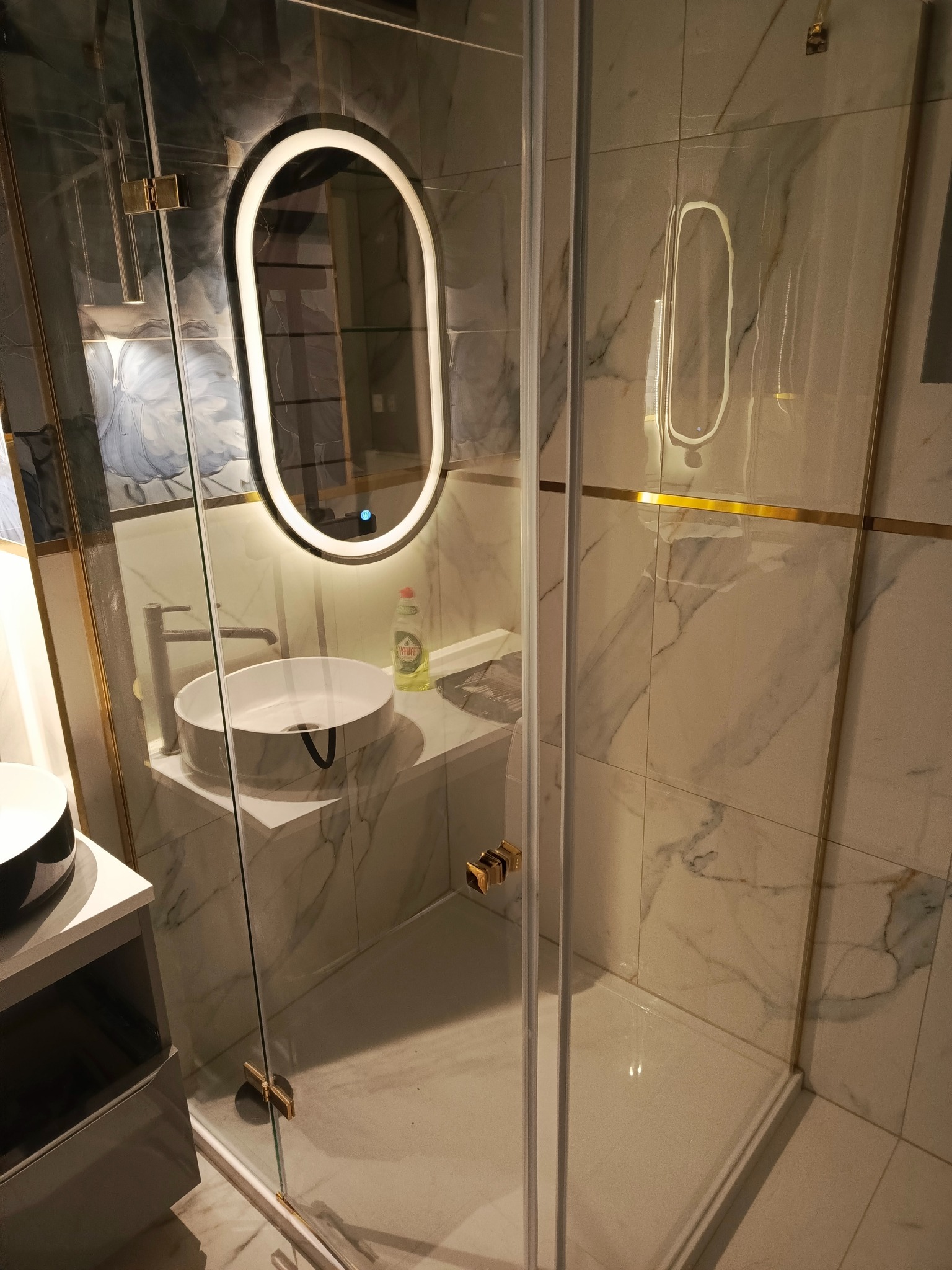 Nowoczesna łazienka z kabiną prysznicową ze złotymi detalami, umywalką nablatową i podświetlanym lustrem owalnym. Marmurowe płytki na ścianach z dekoracyjnymi złotymi paskami. Widoczny płyn...
