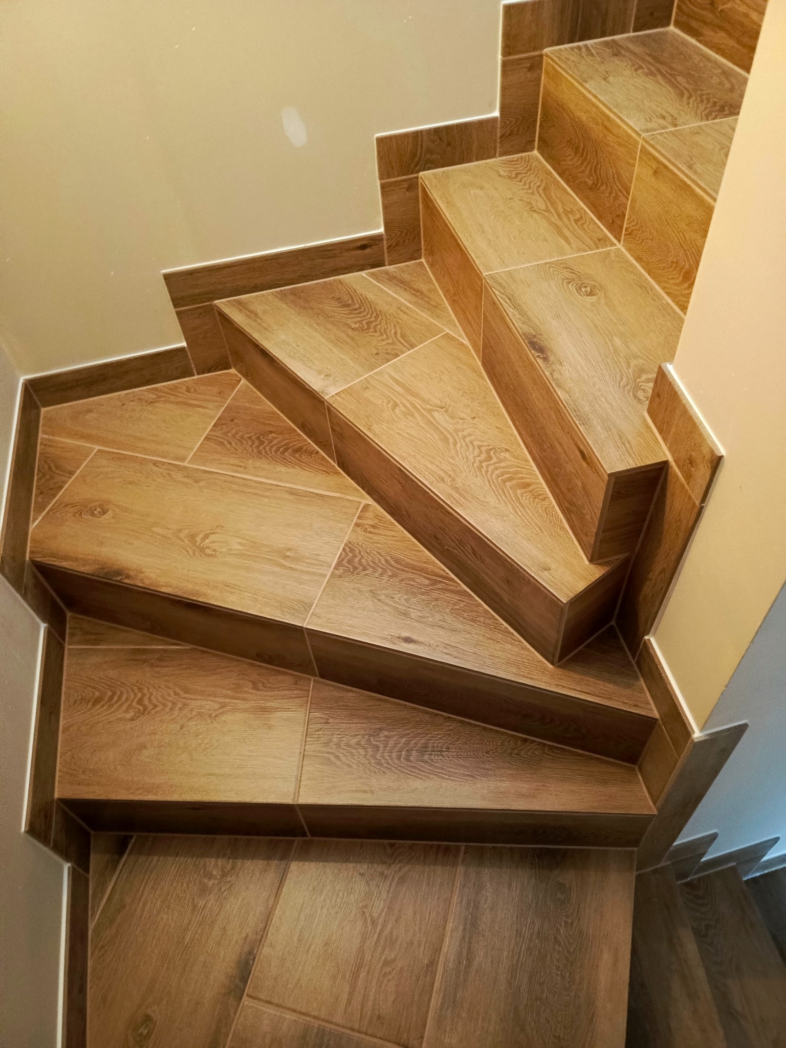 Schody wyłożone płytkami imitującymi drewno, z widocznymi fugami i listwami przypodłogowymi w kolorze płytek, sfotografowane z góry.