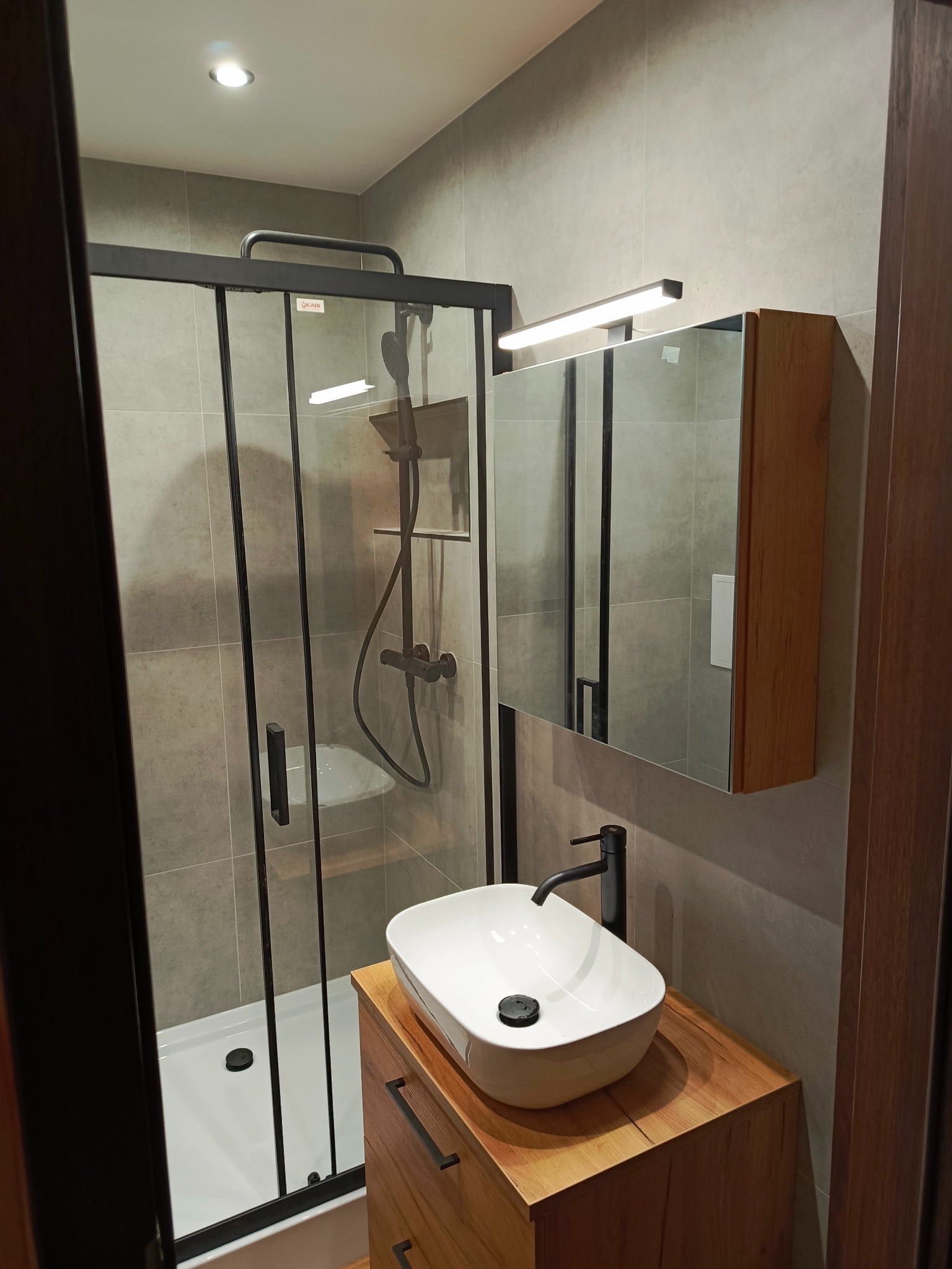Nowoczesna łazienka z kabiną prysznicową typu walk-in z czarnymi profilami, umywalką nablatową w kształcie łzy na drewnianej szafce oraz oświetleniem LED nad lustrem.