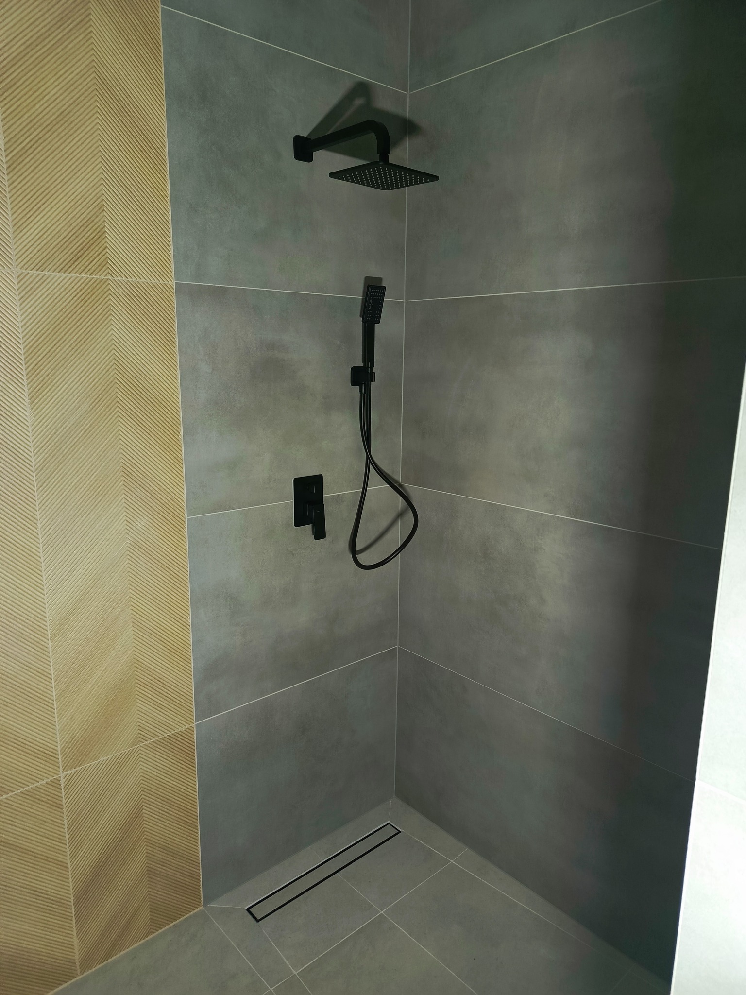 Nowoczesna kabina prysznicowa z szarymi płytkami i akcentem w postaci beżowej ściany z wzorem chevron, czarna armatura, w tym deszczownica i odpływ liniowy.