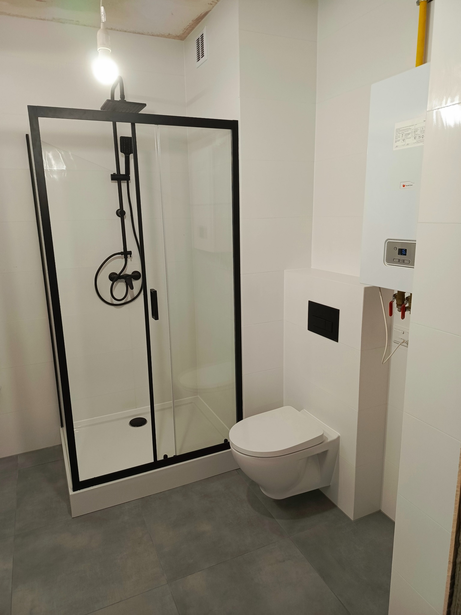 Nowoczesna łazienka z kabiną prysznicową typu walk-in z czarnymi profilami, podwieszaną toaletą z czarnym przyciskiem spłukiwania oraz białym piecem gazowym na ścianie z widocznymi rurami i zaworami.