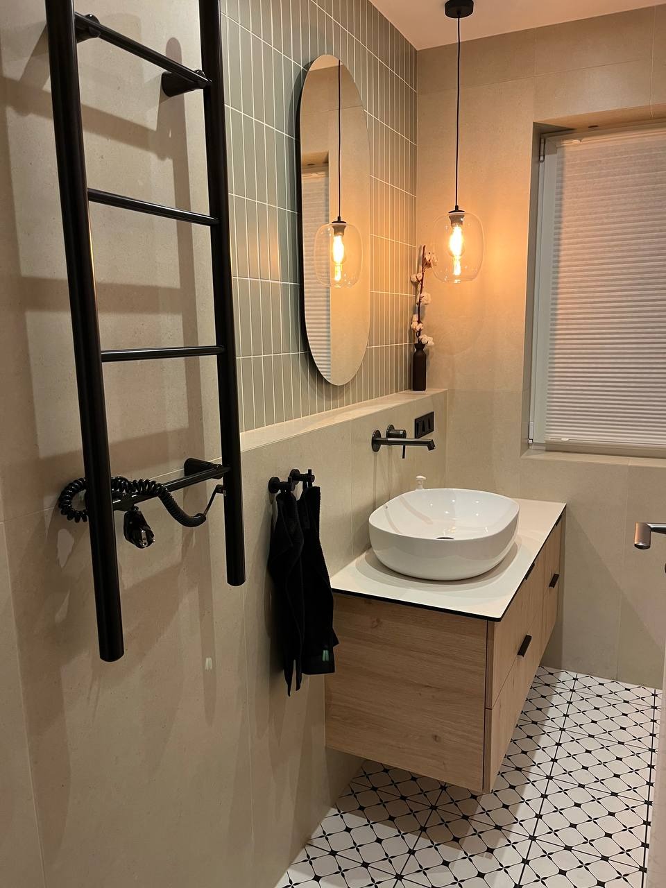Nowoczesna łazienka w beżowej tonacji z owalnym lustrem, wiszącymi lampami z żarówkami Edisona, czarnym grzejnikiem drabinkowym i białą umywalką nablatową na drewnianej szafce, wykończona płytkami...