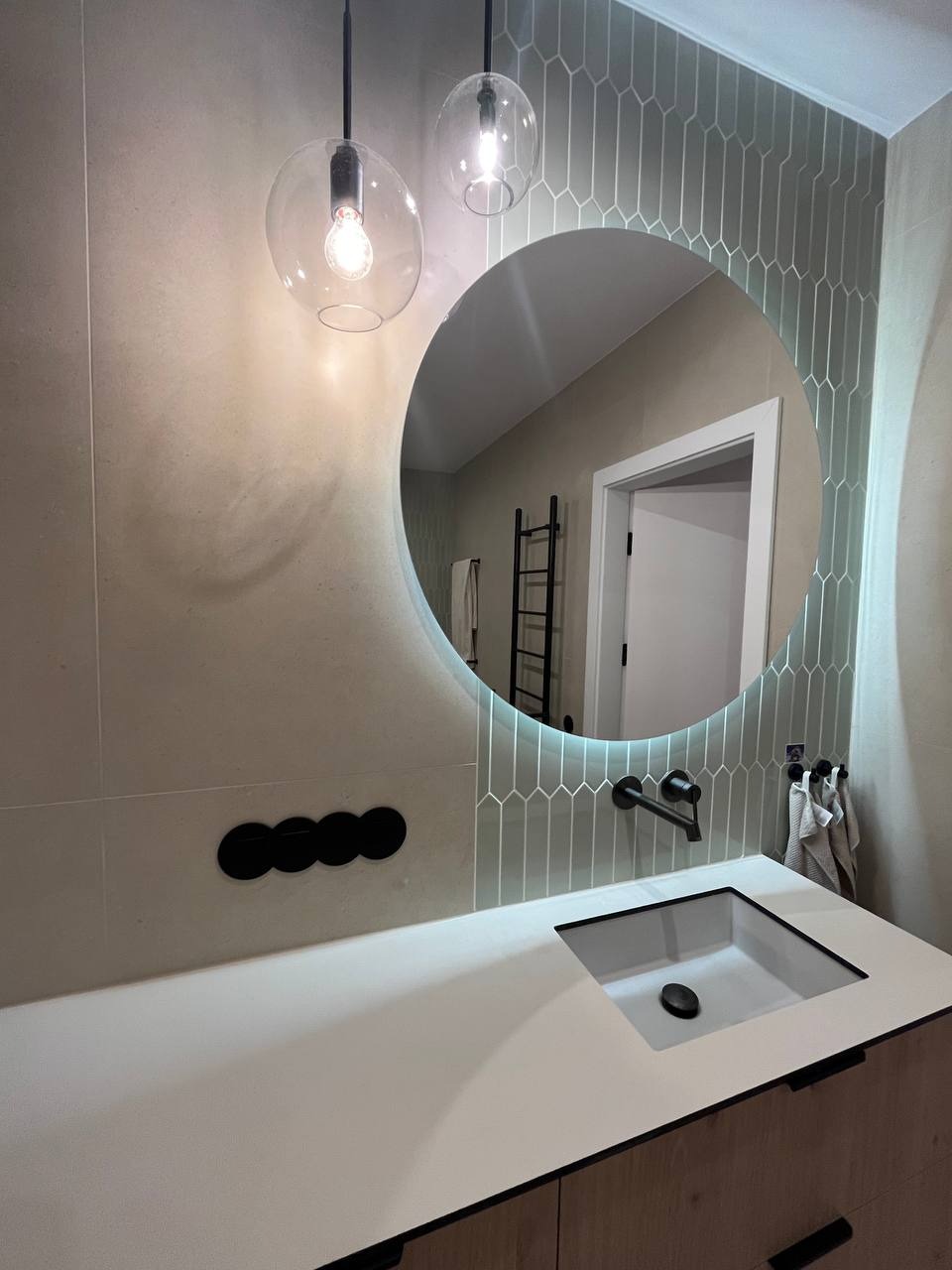Nowoczesna łazienka z okrągłym lustrem z podświetleniem LED, białą umywalką nablatową, czarną armaturą i geometrycznymi płytkami na ścianie, oświetlona wiszącymi lampami z przezroczystego szkła.