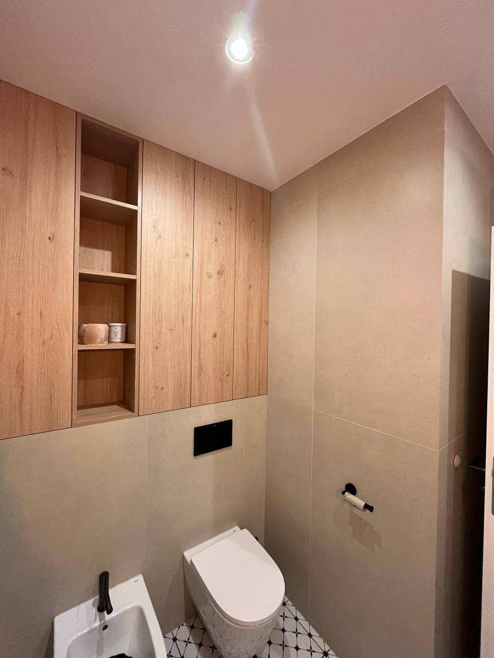 Nowoczesna łazienka w beżowych tonacjach z drewnianymi szafkami wiszącymi, białą toaletą podwieszaną i bidetem na tle płytek z geometrycznym wzorem.