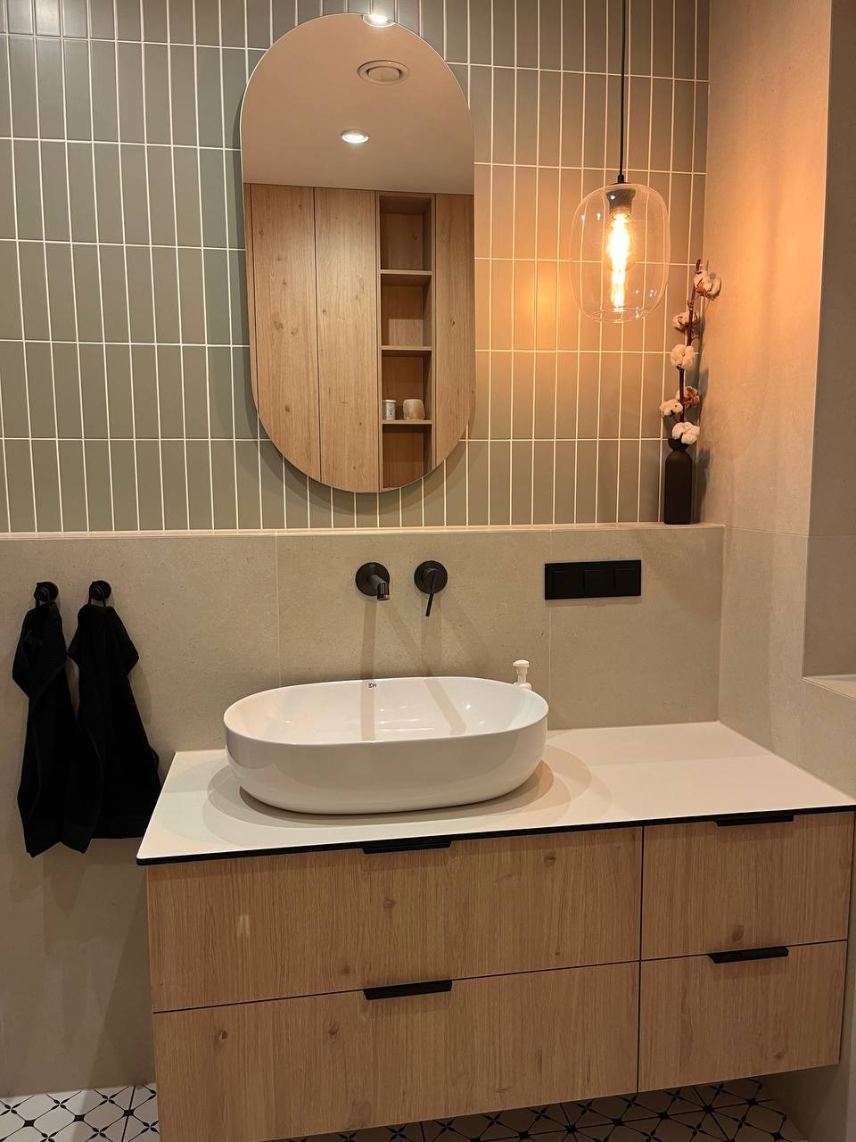 Nowoczesna łazienka z owalną umywalką nablatową, czarną armaturą ścienną i lustrem w drewnianej szafce, oświetlona wiszącą lampą z żarówką Edisona, akcentowana dekoracyjną gałązką w wazonie...