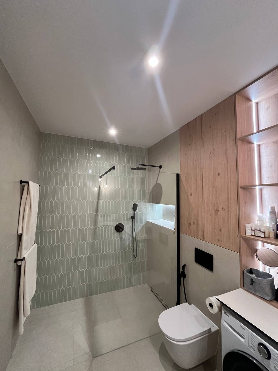 Nowoczesna łazienka z kabiną prysznicową typu walk-in, wyłożona pionowymi, szarymi płytkami w kształcie migdałów, z czarną armaturą i drewnianymi półkami na kosmetyki nad pralką.