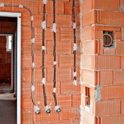 INSTALLATORE - Surowy mur z czerwonej cegły z widocznymi przewodami elektrycznymi w trakcie montażu, prowadzącymi do puszek instalacyjnych; w tle widoczne ościeżnice drzwiowe.
