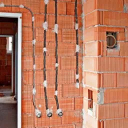 Surowy mur z czerwonej cegły z widocznymi przewodami elektrycznymi w trakcie montażu, prowadzącymi do puszek instalacyjnych; w tle widoczne ościeżnice drzwiowe.