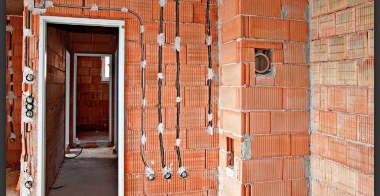 Surowy mur z czerwonej cegły z widocznymi przewodami elektrycznymi w trakcie montażu, prowadzącymi do puszek instalacyjnych; w tle widoczne ościeżnice drzwiowe.