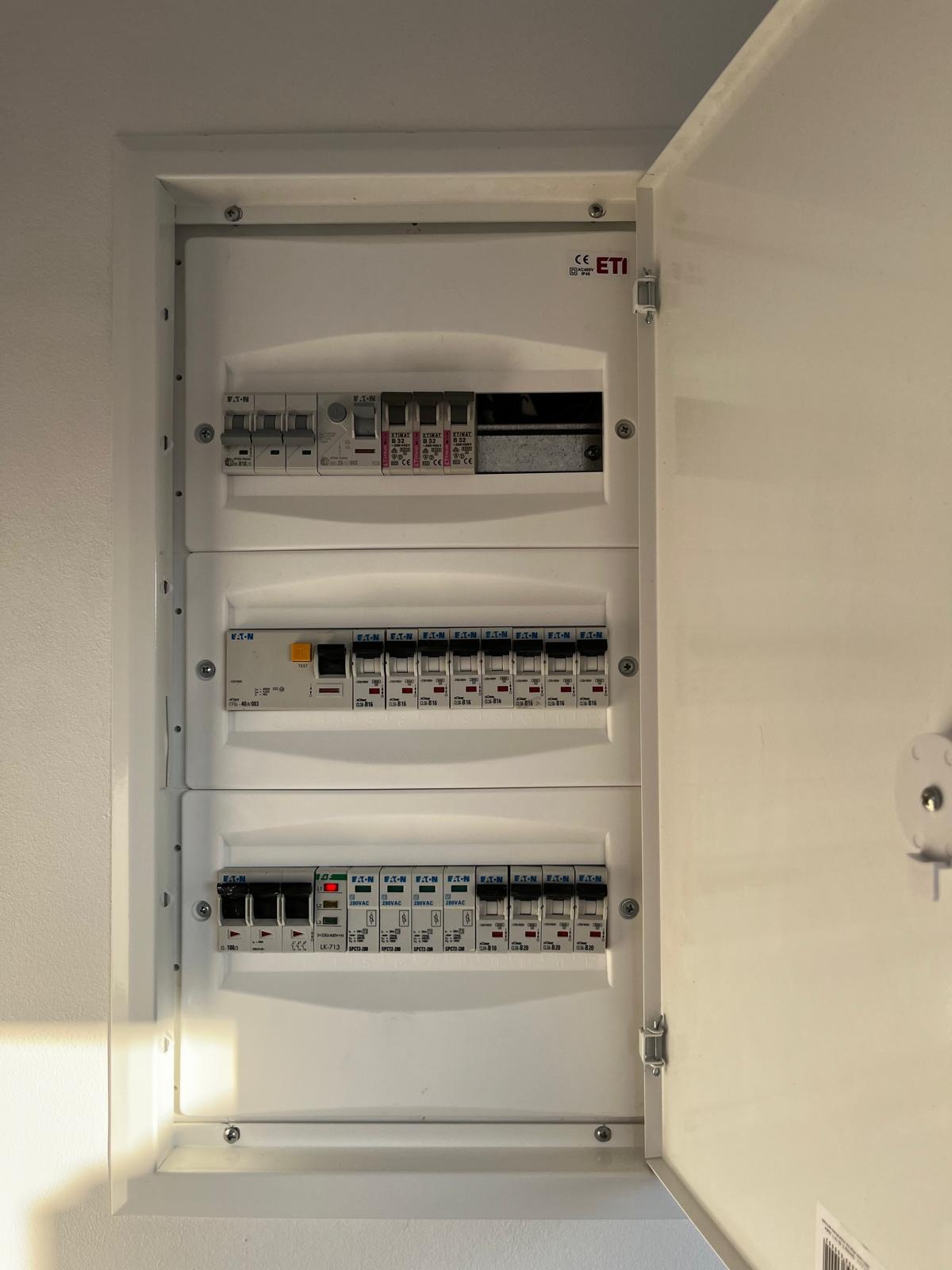 Otwarta skrzynka bezpiecznikowa z widocznymi bezpiecznikami i wyłącznikami, marki ETI i Eaton, zamontowana na białej ścianie.