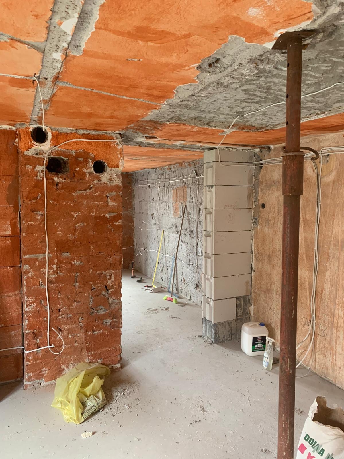 Wnętrze w stanie surowym: widoczne przewody elektryczne wychodzące ze ściany z cegły dziurawki, podwieszony ceglany sufit, wspornik stalowy, białe bloczki betonowe, worki z zaprawą i narzędzia...