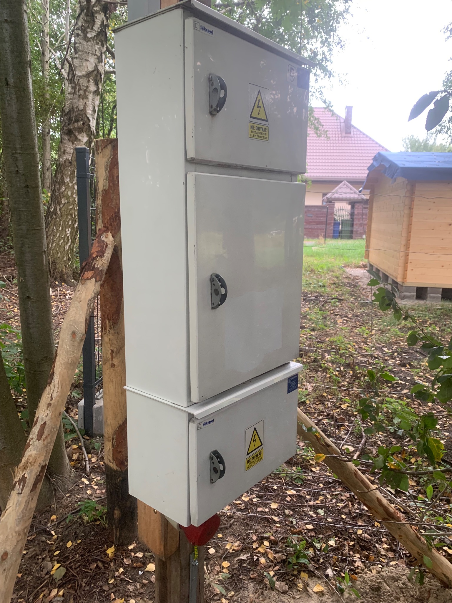 Trzyczęściowa, biała szafka elektryczna z ostrzeżeniami o napięciu, zamontowana na drewnianych słupach na zewnątrz, w tle dom z czerwonym dachem i drewniana altana.