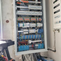 ELGO Instalacje elektryczne Kamil Goch - Rozdzielnica elektryczna w szarej obudowie, z licznymi przewodami i elementami sterującymi. Widoczne narzędzia i fragment ściany w surowym stanie. Przygotowana do Smart Home.