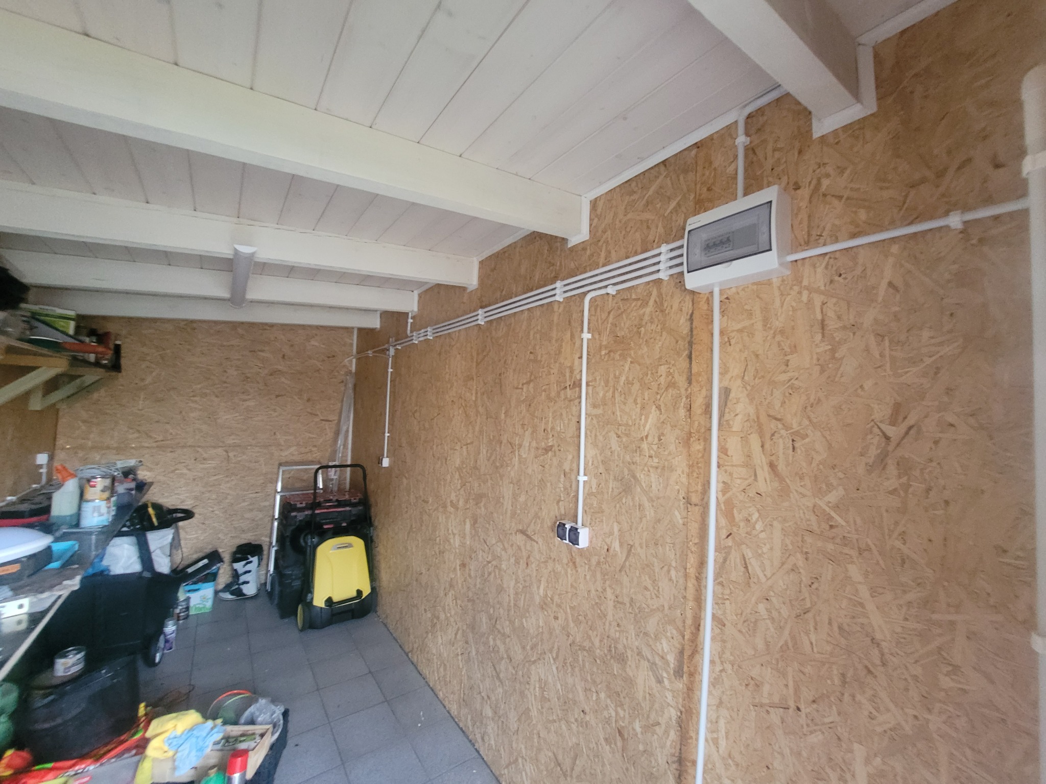 Rozdzielnica elektryczna natynkowa i białe rury elektroinstalacyjne zamocowane na ścianie z płyty OSB w komórce lub garażu, widoczne także gniazdo podwójne i bałagan w tle.