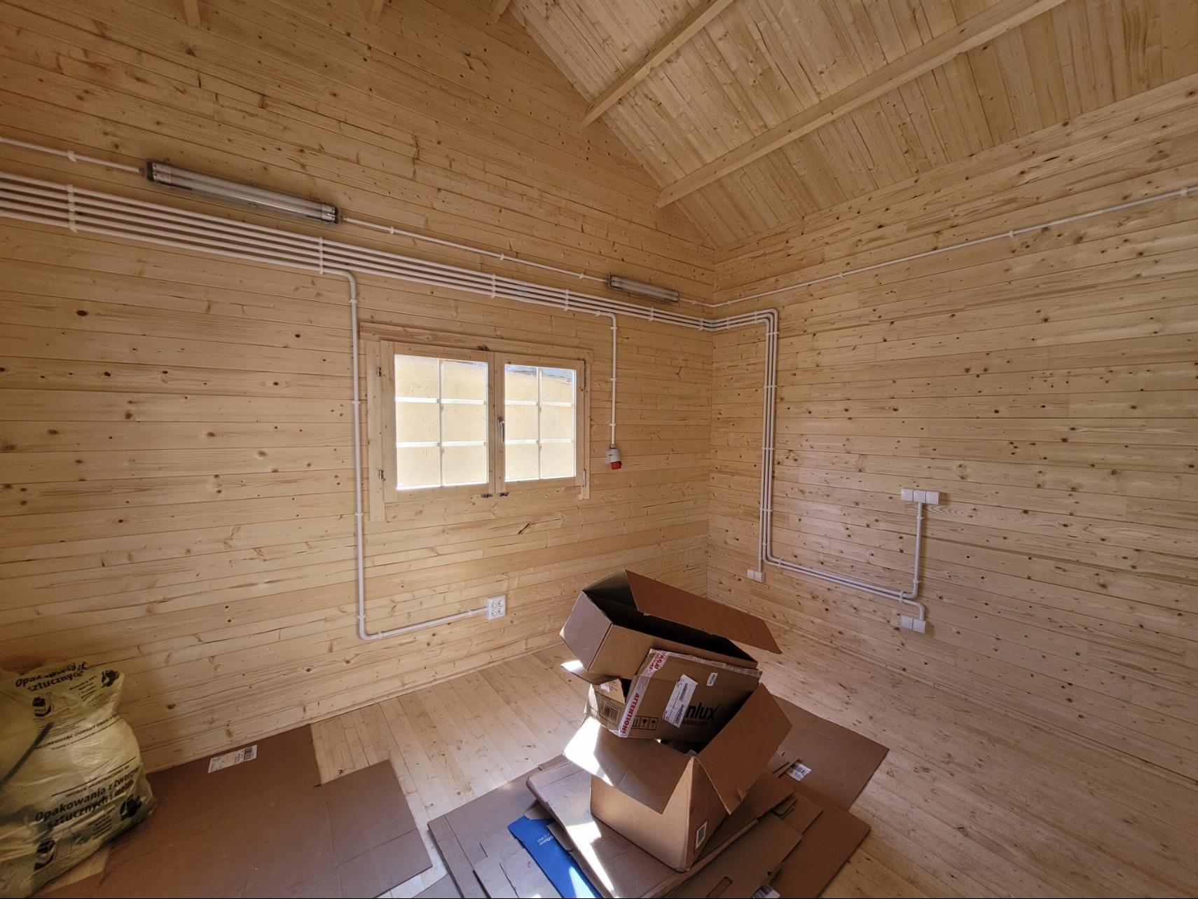 Wnętrze drewnianego domku z widoczną instalacją elektryczną: rurki PCV, lampy, gniazdka. Częściowo rozpakowane kartony na podłodze. Ujęcie szerokokątne.