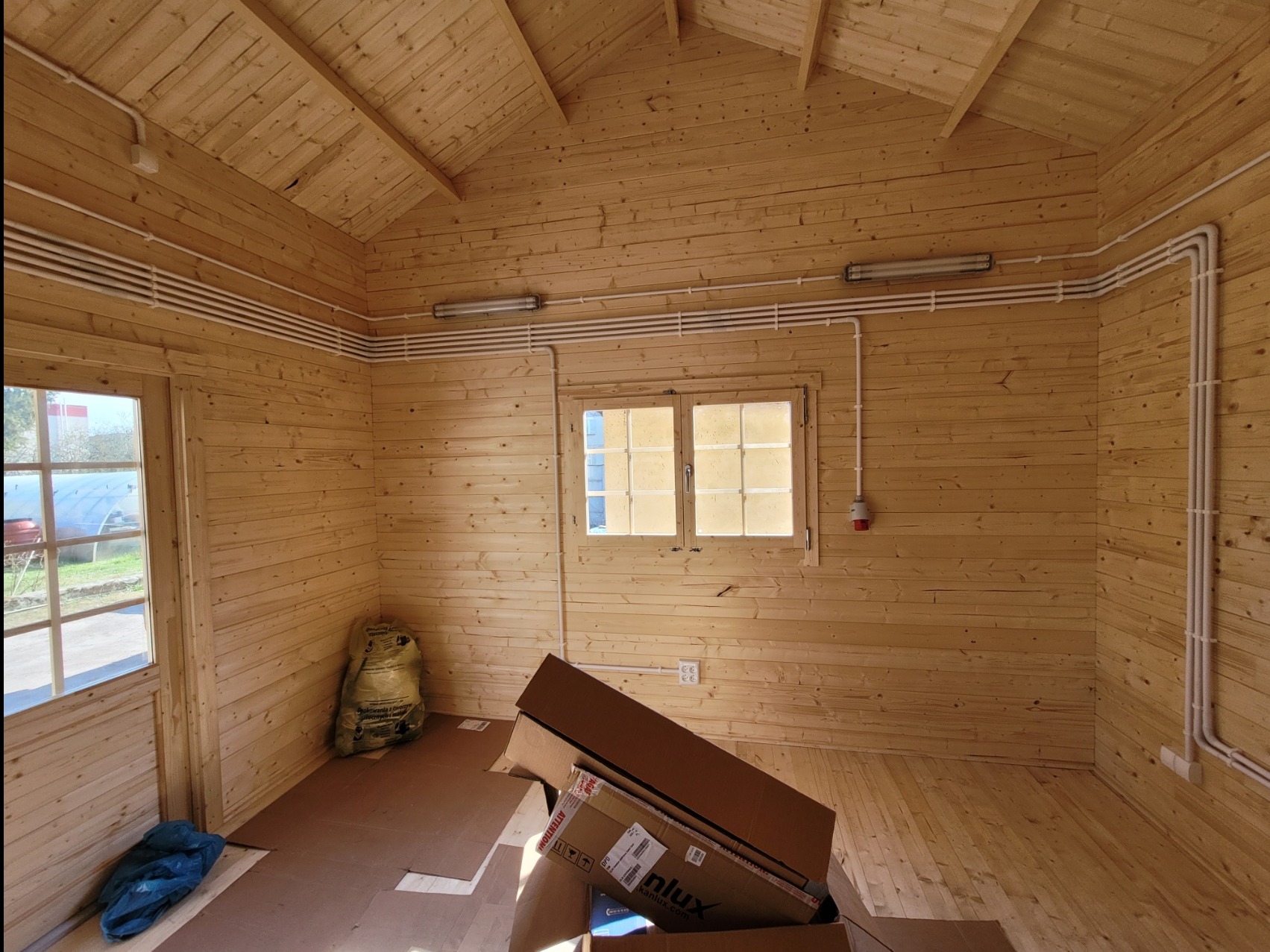 Wnętrze drewnianego domku z widoczną instalacją elektryczną: białe rury na ścianach, lampy jarzeniowe pod sufitem i gniazdo elektryczne przy oknie. Częściowo widoczne kartony na podłodze.