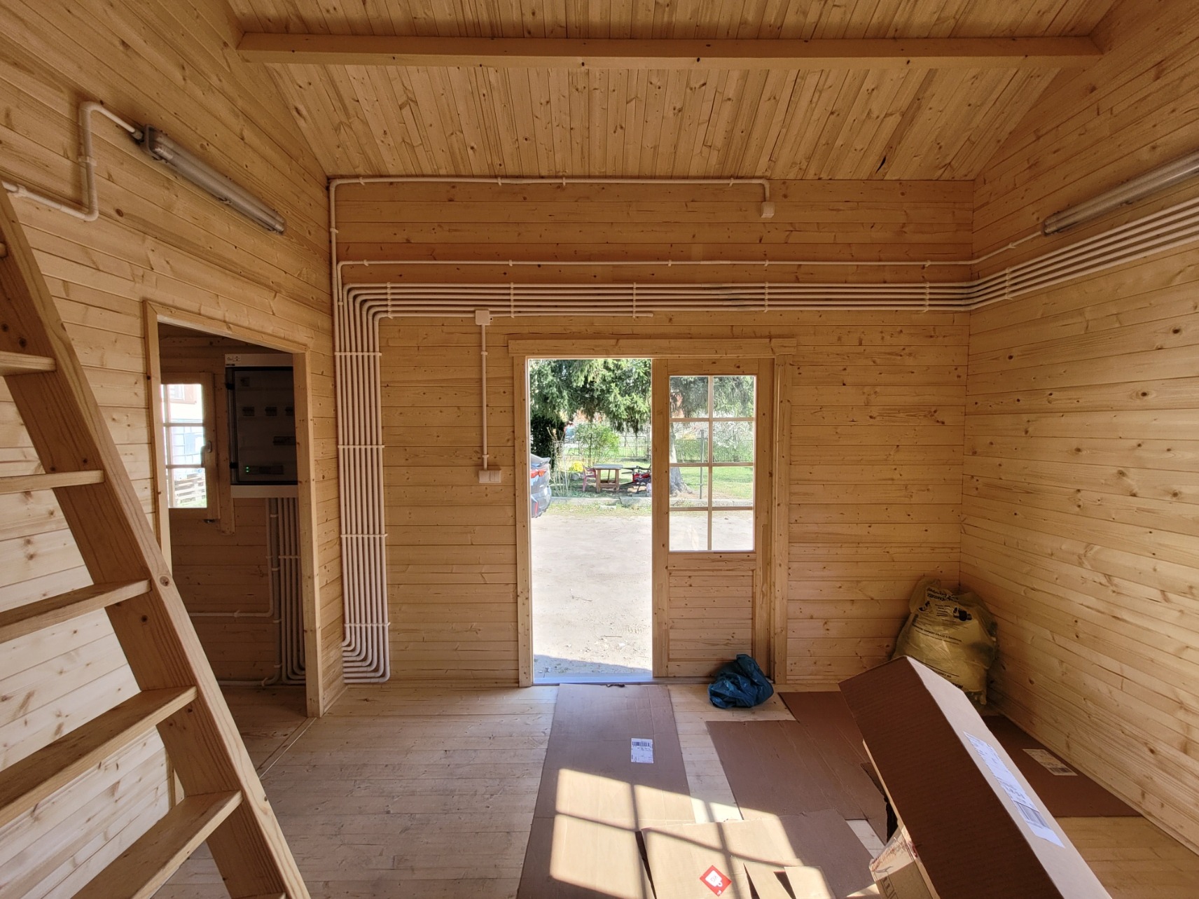 Wnętrze drewnianego domku z widoczną instalacją elektryczną: rurki PCV ułożone na ścianach i suficie, schody drewniane po lewej. Widok na zewnątrz przez przeszklone drzwi.