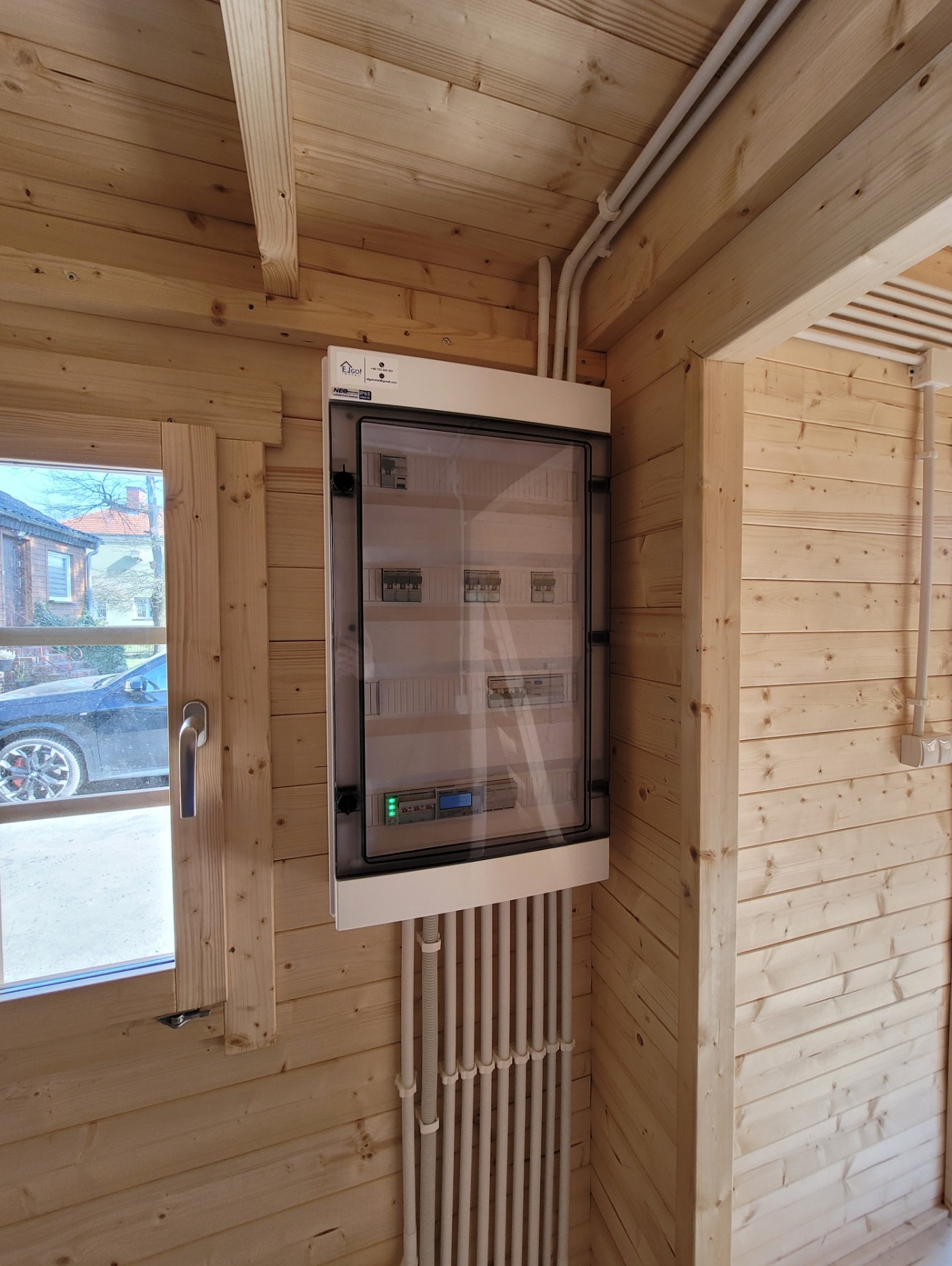 Rozdzielnica elektryczna z przezroczystymi drzwiami, zamontowana w drewnianym domku, widoczne kable i okno z widokiem na samochód.