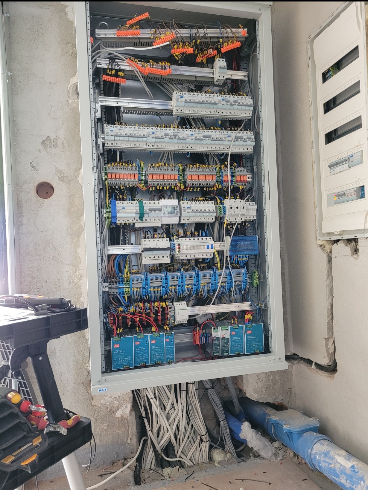 Rozdzielnica elektryczna w szarej obudowie, z licznymi przewodami i elementami sterującymi. Widoczne narzędzia i fragment ściany w surowym stanie. Przygotowana do Smart Home.