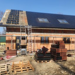 MAR - DEK USŁUGI DEKARSKIE MARCIN CYGAN - Budowa dachu na domu jednorodzinnym: czarne dachówki ułożone częściowo, reszta w pakietach na ziemi, widoczna więźba dachowa i rusztowanie.