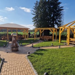 MAR - DEK USŁUGI DEKARSKIE MARCIN CYGAN - Trzy altany ogrodowe z drewnianą konstrukcją, brukowana ścieżka z oświetleniem, dekoracyjny drewniany wiatrak, trawnik i drzewa w tle, słoneczny dzień.