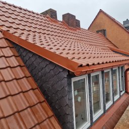 MAR - DEK USŁUGI DEKARSKIE MARCIN CYGAN - Czerwona dachówka ceramiczna na skośnym dachu z widocznymi kominami, obróbkami blacharskimi w kolorze dachówki i fragmentem elewacji pokrytej łupkiem, widok z rusztowania.