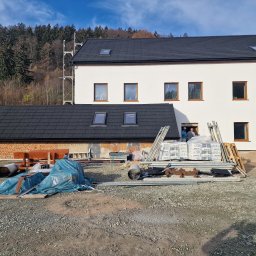 MAR - DEK USŁUGI DEKARSKIE MARCIN CYGAN - Nowy, dwukondygnacyjny budynek z ciemnym dachem z dachówki bitumicznej i białymi ścianami, na placu budowy widoczne materiały budowlane i dwóch pracowników w drzwiach.