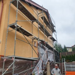 MAR - DEK USŁUGI DEKARSKIE MARCIN CYGAN - Dom jednorodzinny w trakcie prac elewacyjnych, osłonięty folią i otoczony rusztowaniem, z widocznym pracownikiem w bluzie.