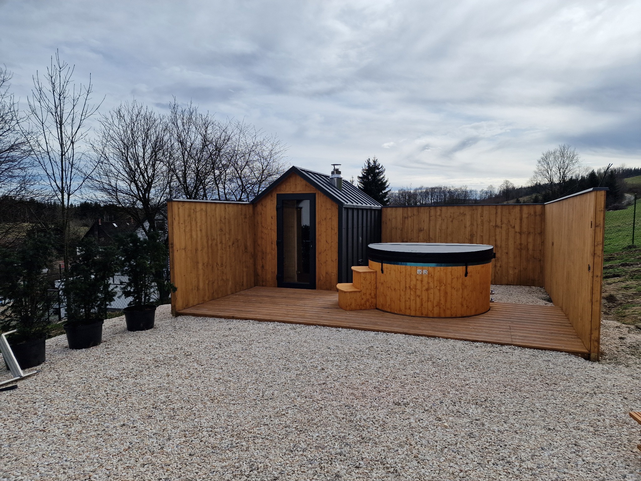 Drewniany taras z jacuzzi i domkiem narzędziowym, otoczony żwirowym podłożem, na tle pochmurnego nieba i drzew bez liści.