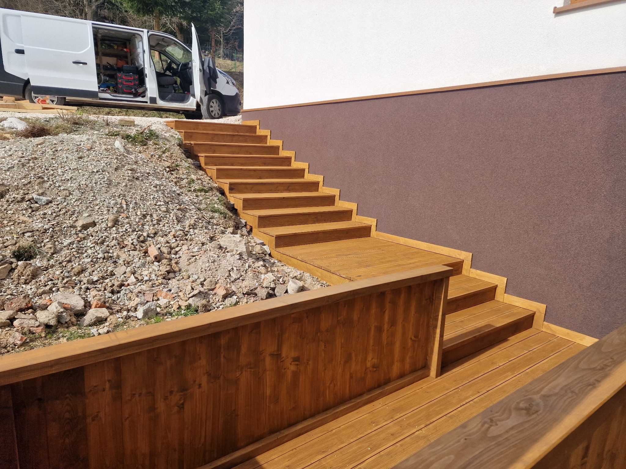 Drewniane schody zewnętrzne prowadzące do domu z brązową elewacją, częściowo ukończone, z widocznym samochodem dostawczym z otwartymi drzwiami i narzędziami w tle.