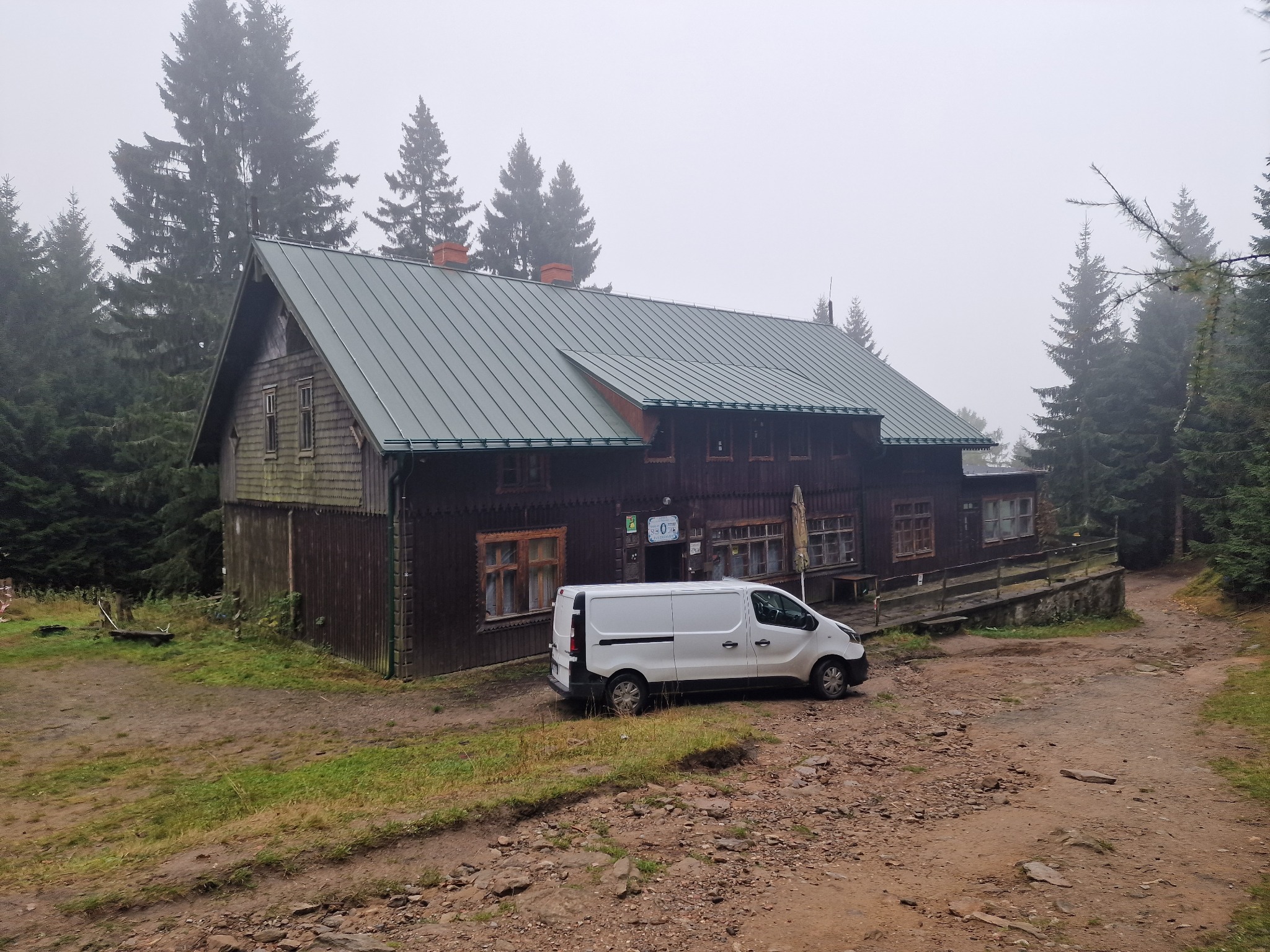 Drewniany budynek w górach z nowym, zielonym dachem blaszanym, widoczny biały samochód dostawczy na szutrowej drodze, mgła w tle.