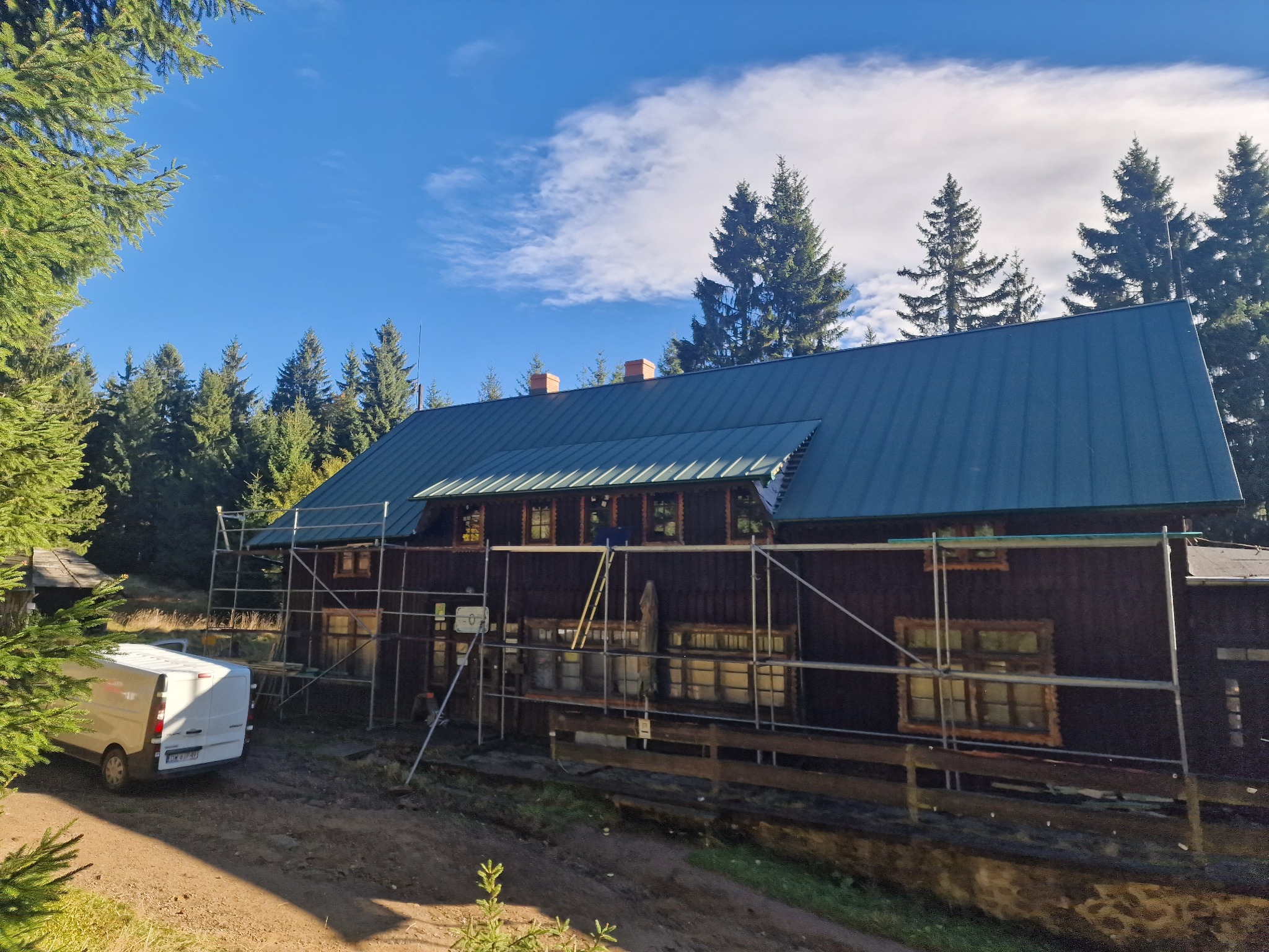 Drewniany budynek z nowym, błyszczącym, niebiesko-zielonym dachem, otoczony rusztowaniami, z białym vanem zaparkowanym obok, na tle lasu i błękitnego nieba z chmurami.