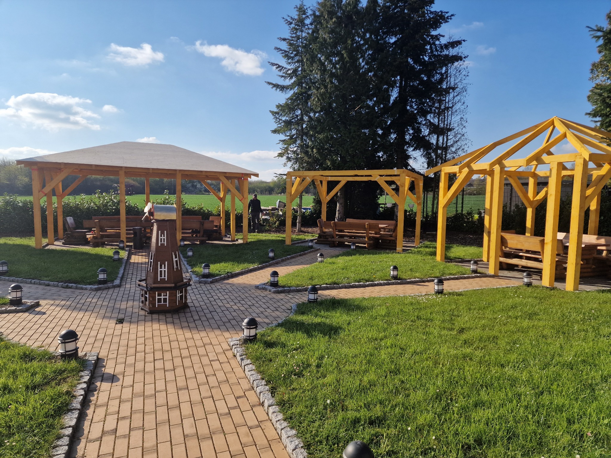 Trzy altany ogrodowe z drewnianą konstrukcją, brukowana ścieżka z oświetleniem, dekoracyjny drewniany wiatrak, trawnik i drzewa w tle, słoneczny dzień.
