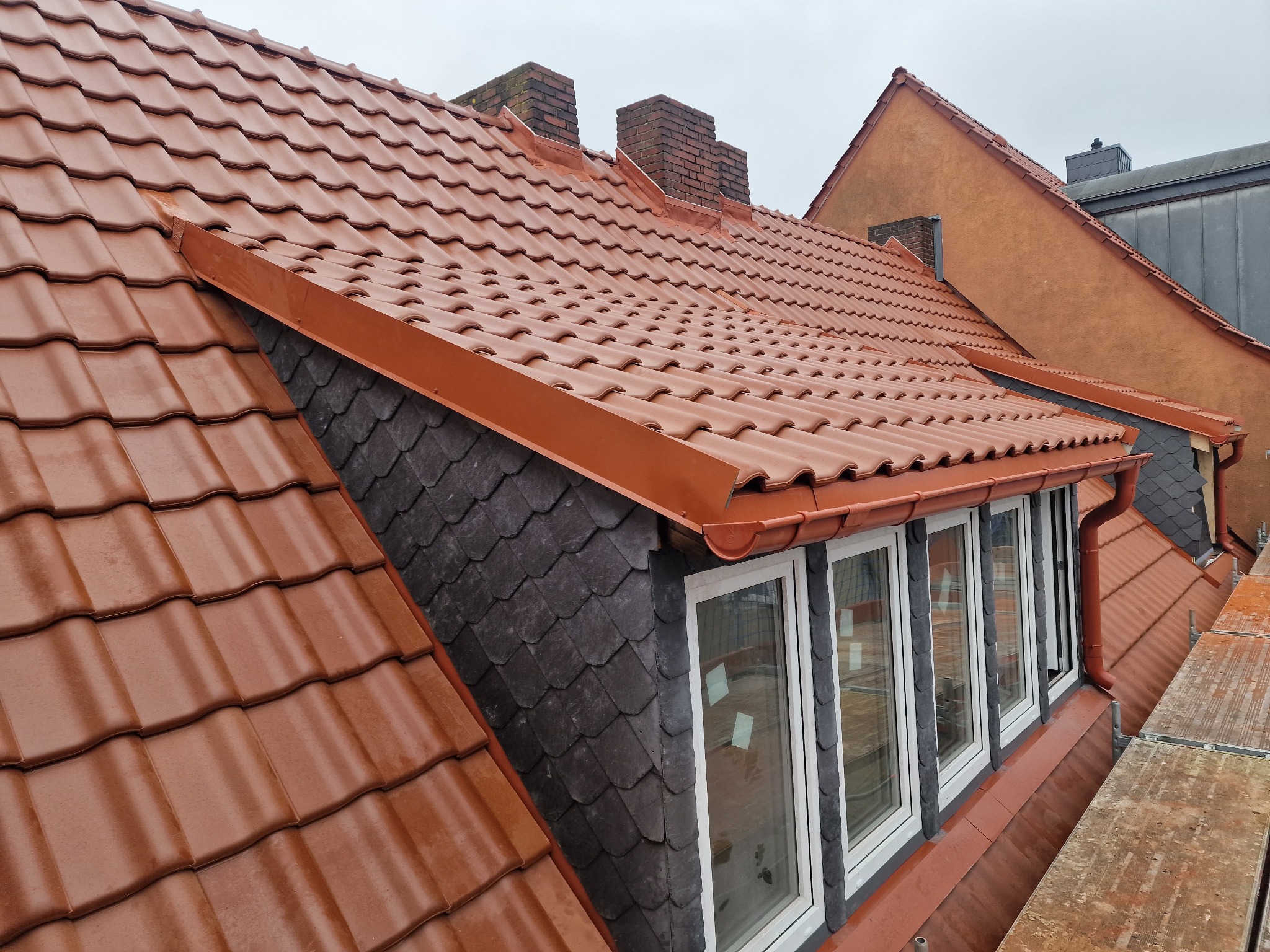 Czerwona dachówka ceramiczna na skośnym dachu z widocznymi kominami, obróbkami blacharskimi w kolorze dachówki i fragmentem elewacji pokrytej łupkiem, widok z rusztowania.