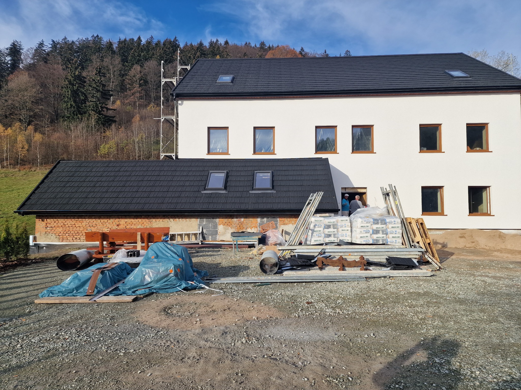 Nowy, dwukondygnacyjny budynek z ciemnym dachem z dachówki bitumicznej i białymi ścianami, na placu budowy widoczne materiały budowlane i dwóch pracowników w drzwiach.