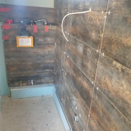 Konrad Ciepliński - Układanie ciemnych płytek imitujących drewno w łazience, widoczne krzyżyki dystansowe, poziomica, wiertarka akumulatorowa i instrukcja obsługi na ścianie z płyt gipsowo-kartonowych.