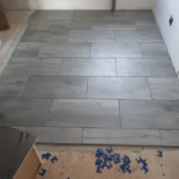 Układanie szarych płytek podłogowych imitujących drewno w pomieszczeniu, widoczne niebieskie klipsy do poziomowania płytek i rura kanalizacyjna przy ścianie.