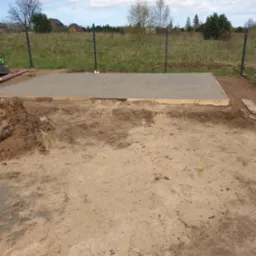 Wylana betonowa płyta fundamentowa z drewnianym szalunkiem na piaszczystym terenie działki, widoczna siatka ogrodzeniowa i zieleń w tle.