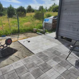 Układanie szarej kostki brukowej o wzorze drewna na podsypce piaskowej przy drewnianym budynku, widoczny wózek z narzędziami i poziomica.