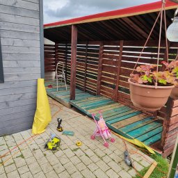 Konrad Ciepliński - Budowa altany z drewnianą konstrukcją i czerwonym dachem. Widoczne narzędzia, zabawkowy wózek i donica z kwiatami. Częściowo ułożona kostka brukowa.