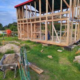 Konrad Ciepliński - Szkielet drewnianej konstrukcji budynku z czerwonym dachem w trakcie budowy, widoczne narzędzia i betoniarka w tle, na trawiastym terenie.