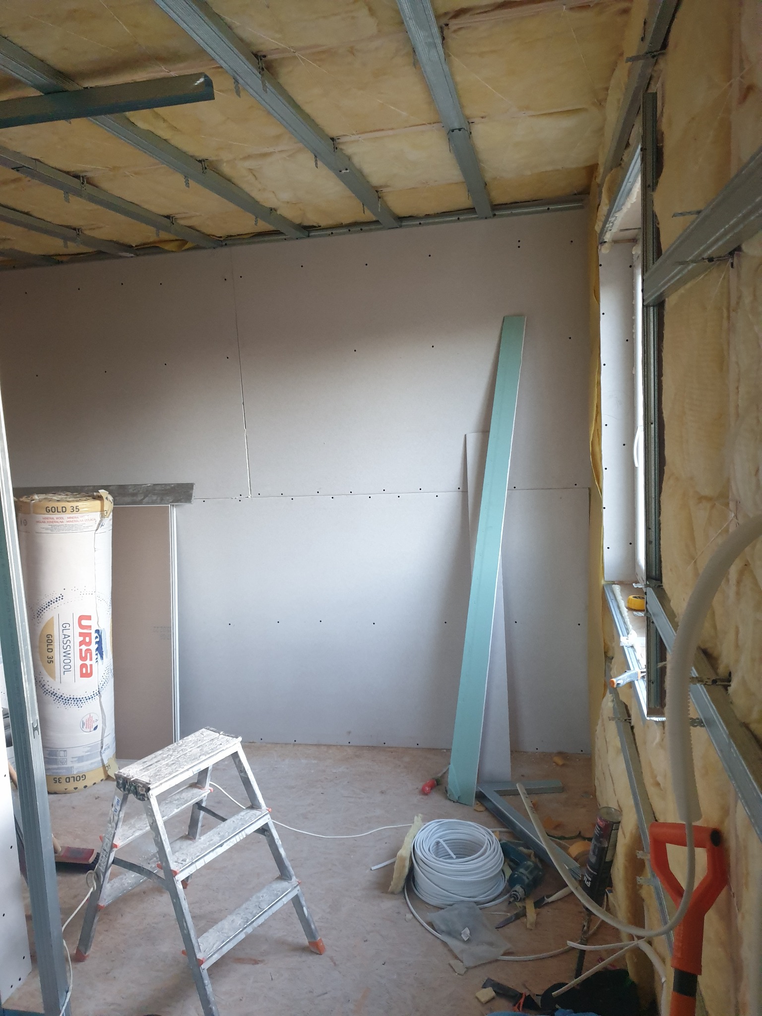 Wnętrze w trakcie prac wykończeniowych: ściany obłożone płytami kartonowo-gipsowymi, izolacja z wełny mineralnej na suficie i ścianach, metalowe profile, drabina, rolka wełny Ursa Gold 35...