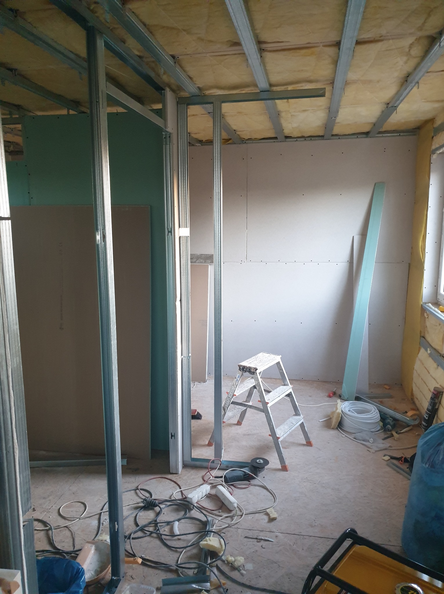 Montaż metalowej konstrukcji pod płyty karton-gips w trakcie remontu, widoczne profile, drabina, przewody i narzędzia na podłodze.