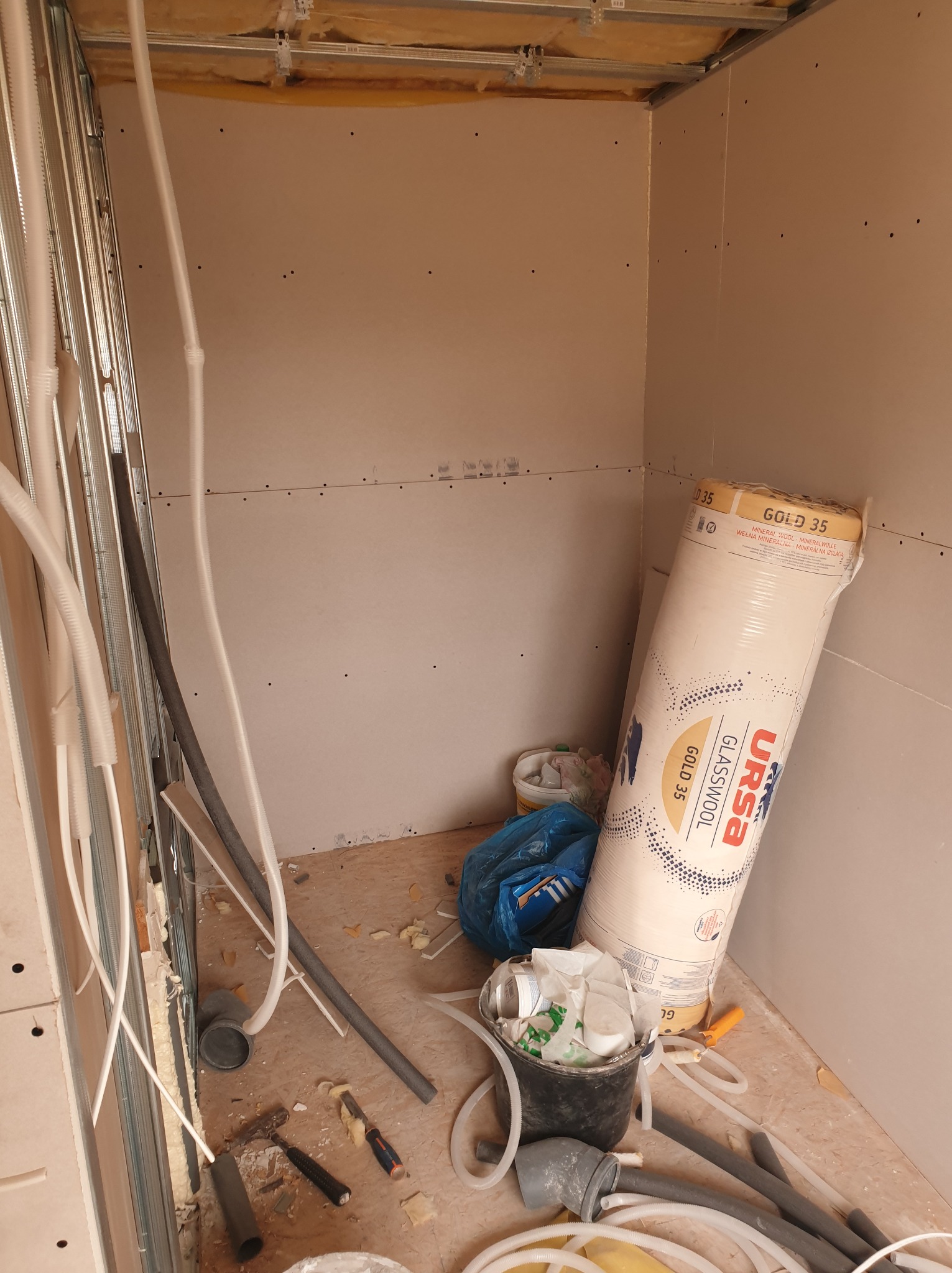 Wnętrze w trakcie prac wykończeniowych: widoczne profile metalowe, instalacje elektryczne, płyty gipsowo-kartonowe oraz rolka wełny mineralnej URSA Gold 35, a także narzędzia i odpady budowlane.
