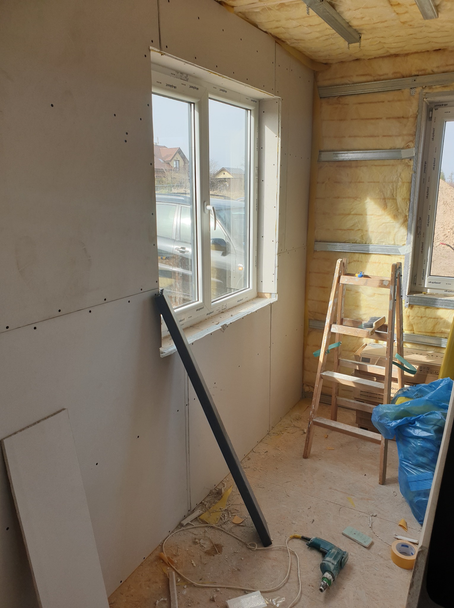 Wnętrze w trakcie prac wykończeniowych: ściany pokryte płytami kartonowo-gipsowymi, widoczne okno, drabina oparta o ścianę z izolacją z wełny mineralnej, narzędzia na podłodze, wiertarka i taśma...